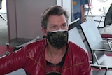 A man in a red jacket wears a mask while pictured seated in an airport lounge