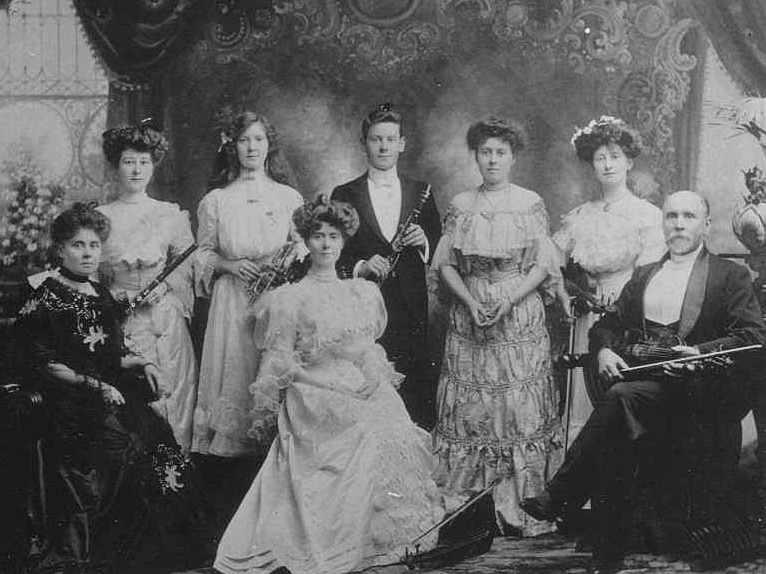 A group of people in1900 clothing holding musical instruments pose for a postcard.