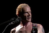 Red Hot Chili Peppers bass player Flea stands shirtless in front of a microphone, with a guitar strapped to him