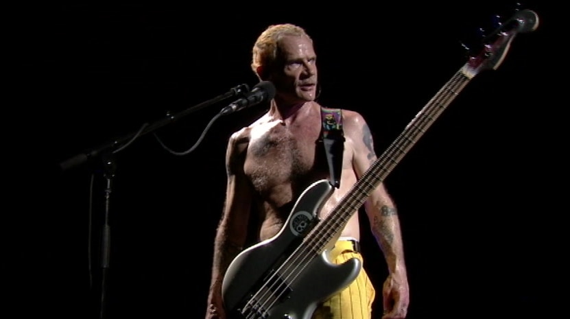 Red Hot Chili Peppers bass player Flea stands shirtless in front of a microphone, with a guitar strapped to him