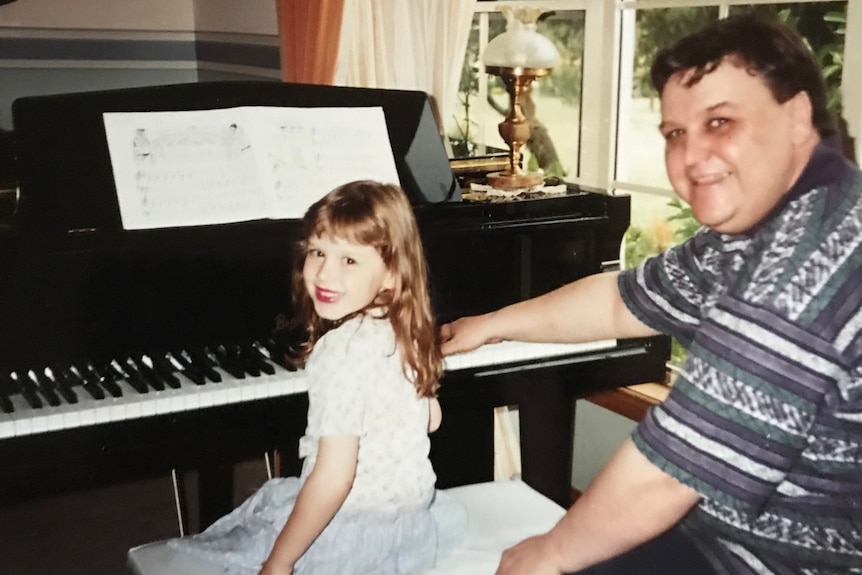 A brunette 7-year-old girl sits at a piano, smiling, beside her dad, a middle-aged white man with greying dark hair.