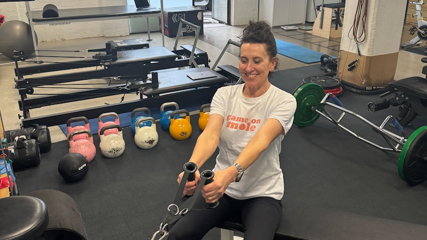 Virginia Trioli pulls a weight machine towards her as she sits, smiling, in workout gear