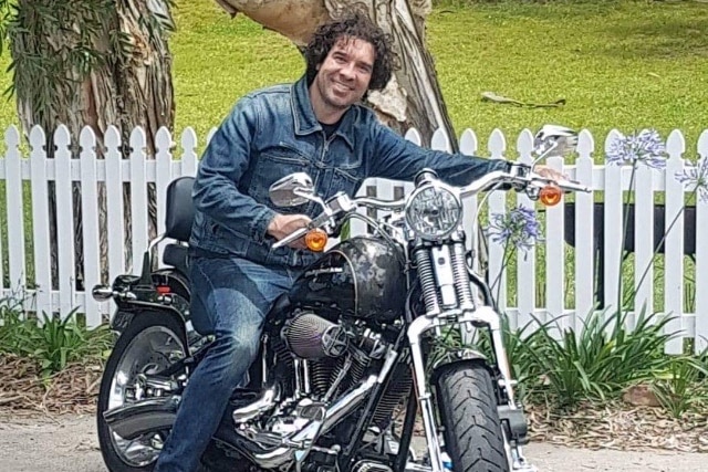 Shane has curly brown hair and is wearing a denim jacket and denim pants sitting on a motocycle and smiling at the camera.