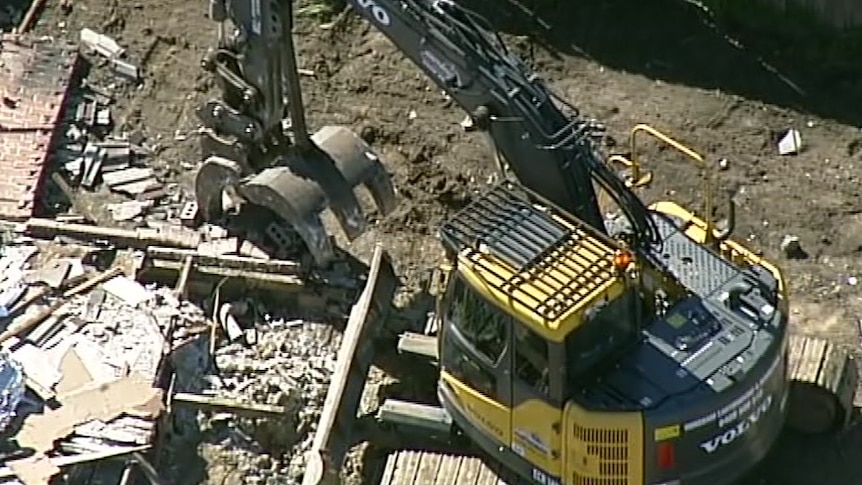 A large digger sits amongst the rubble of the home, including a collapsed brick wall.