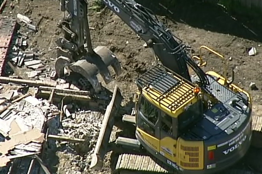 A large digger sits amongst the rubble of the home, including a collapsed brick wall.