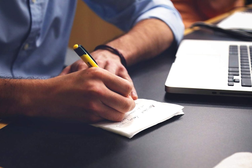 A man is writing on a notepad and using a laptop.