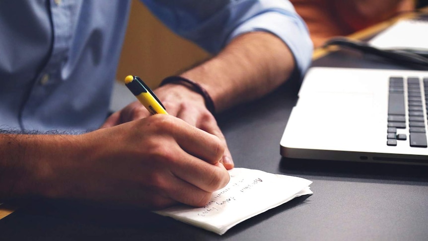 A man is writing on a notepad and using a laptop.