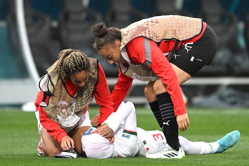 A woman footballer wearing white lays on the grass with her face in her hands as two team-mates in bibs comfort her