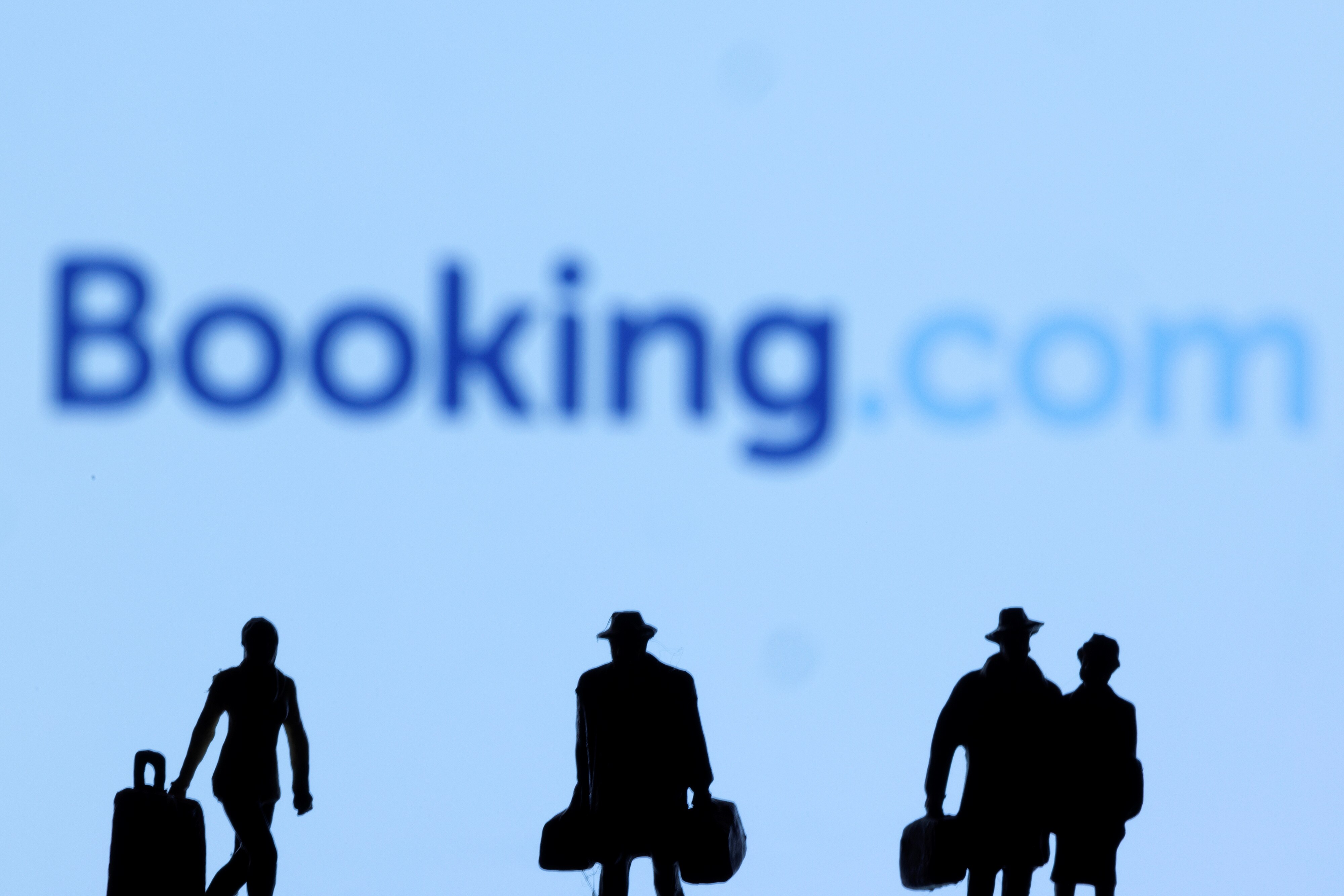 Figurines of travellers walking with bags, in front of a large Booking.com logo in a photographer's illustration.