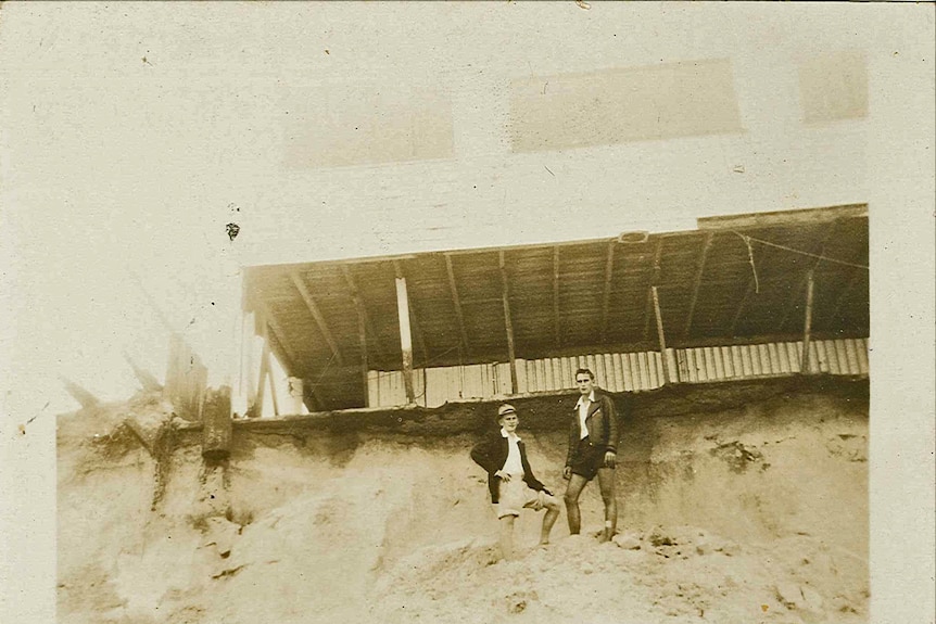 Two men stand under a house overhanging sand dunes on Main Beach, Gold Coast, February 20, 1954