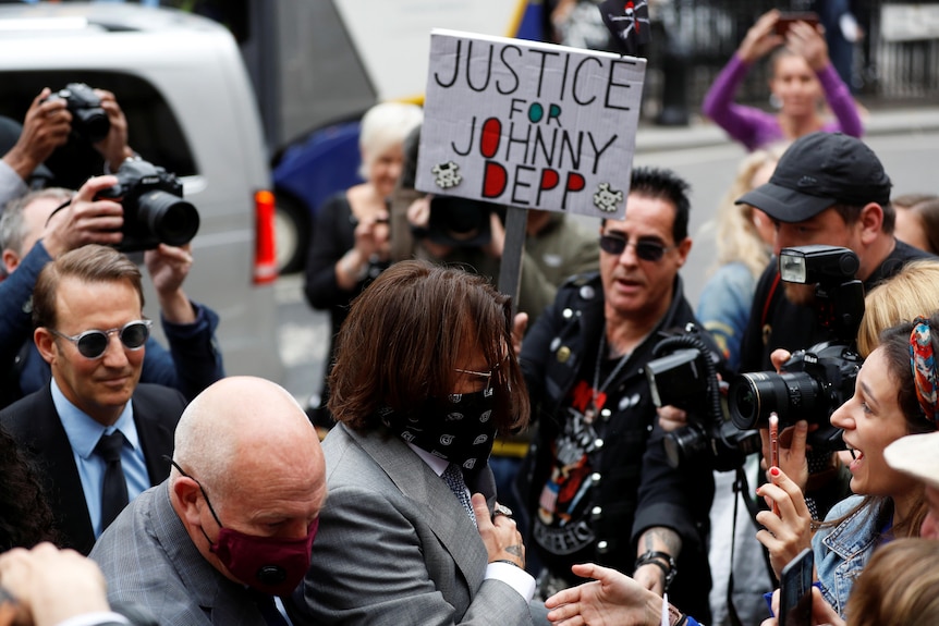 Johnny Depp in a grey suit is escorted through a crowd of photographers and fans, one holding a JUSTICE FOR JOHNNY DEPP sign