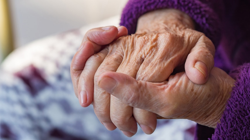 An elderly person holds their hands together in their lap.