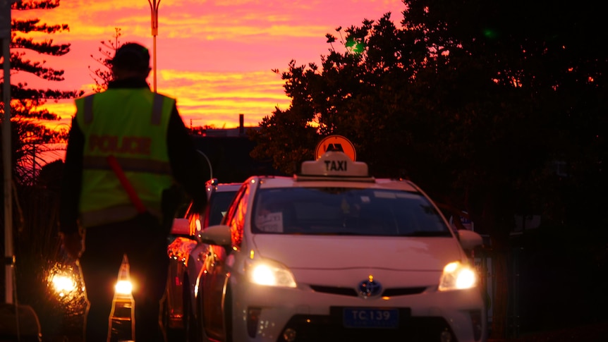 A queue of taxis seen sunrise, with the rear view of a police officer in hi-viz vest.