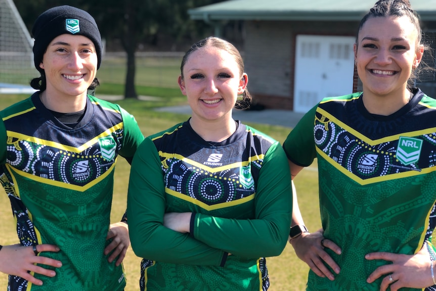The three athletes stand in a row wearing green jerseys and smiling, outside.