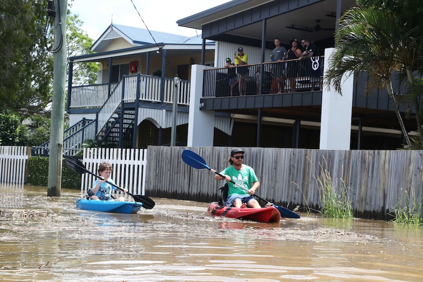 People stand on a verandah watching as a man and a child paddle a kayak down a street.