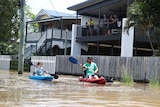 People stand on a verandah watching as a man and a child paddle a kayak down a street.