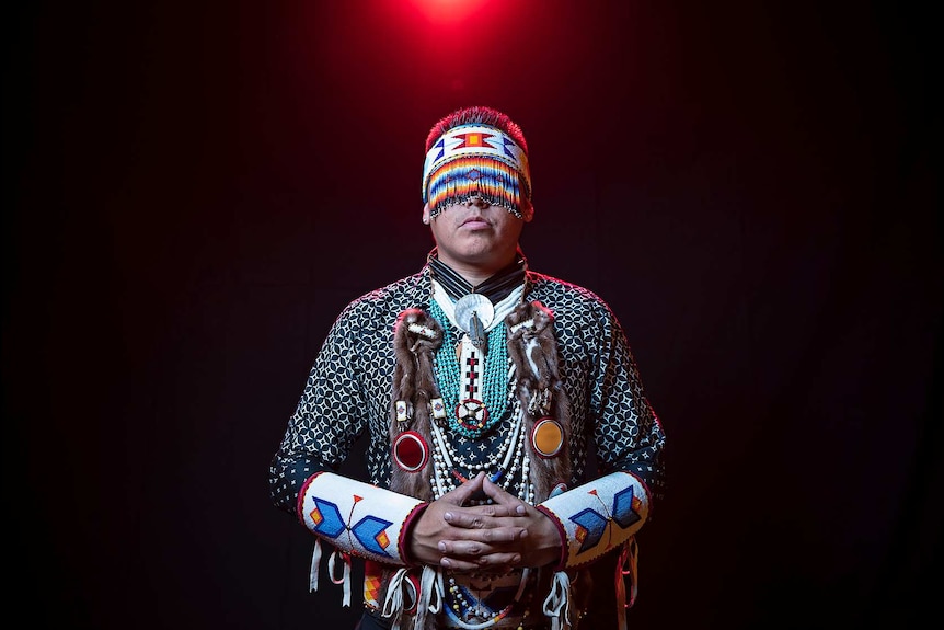 A man with beaded head piece covering eyes wears native American regalia in front of black backdrop with small red light above.