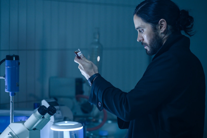 A long-haired, middle-aged man stands in a scientific lab holding a vial of blood