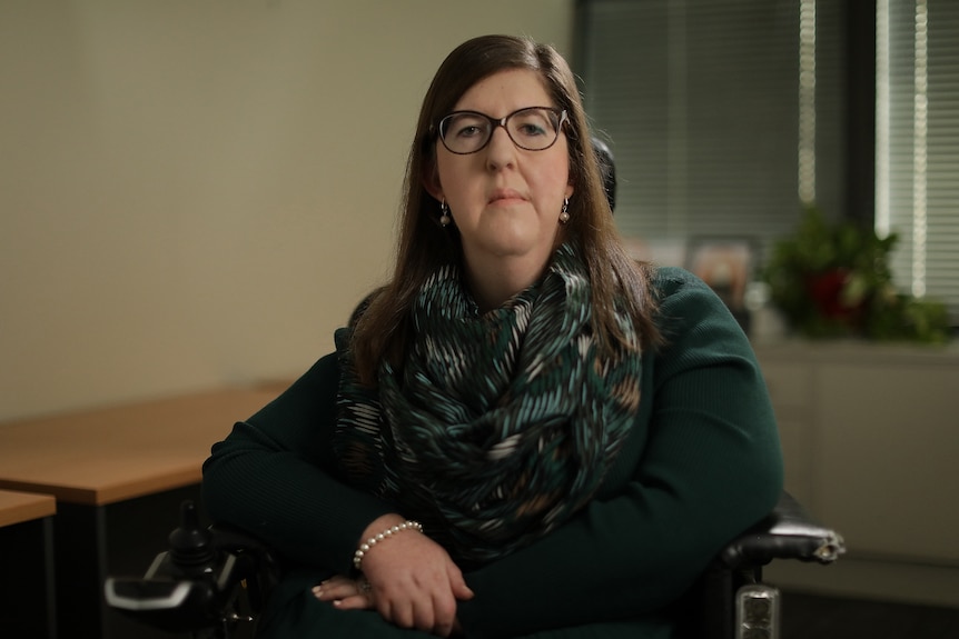 A woman wearing glasses, using an electric wheelchair, sitting in an office looks at the camera with a serious expression.