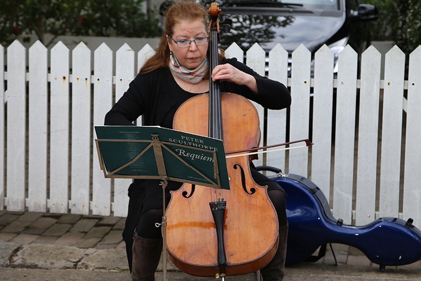 A cellist with red hair performs in front of a white picket fence.