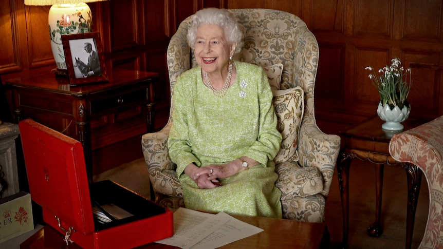 The Queen sits on a lounge chair with an open red box on a table in front of her.