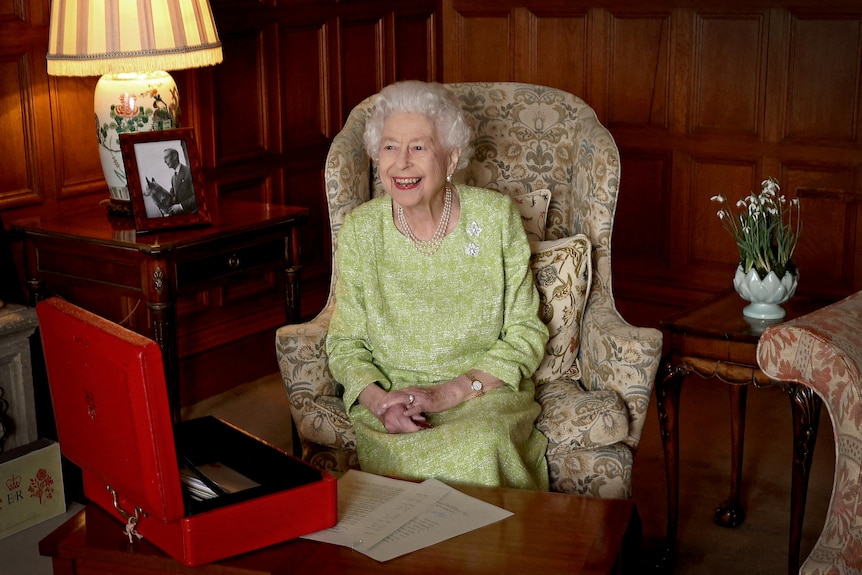 The Queen sits on a lounge chair with an open red box on a table in front of her.