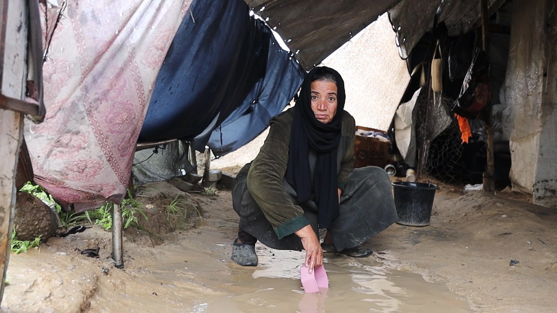 A woman wearing a hijab crouching under a tent on top of a puddle using a plastic item to scoop water