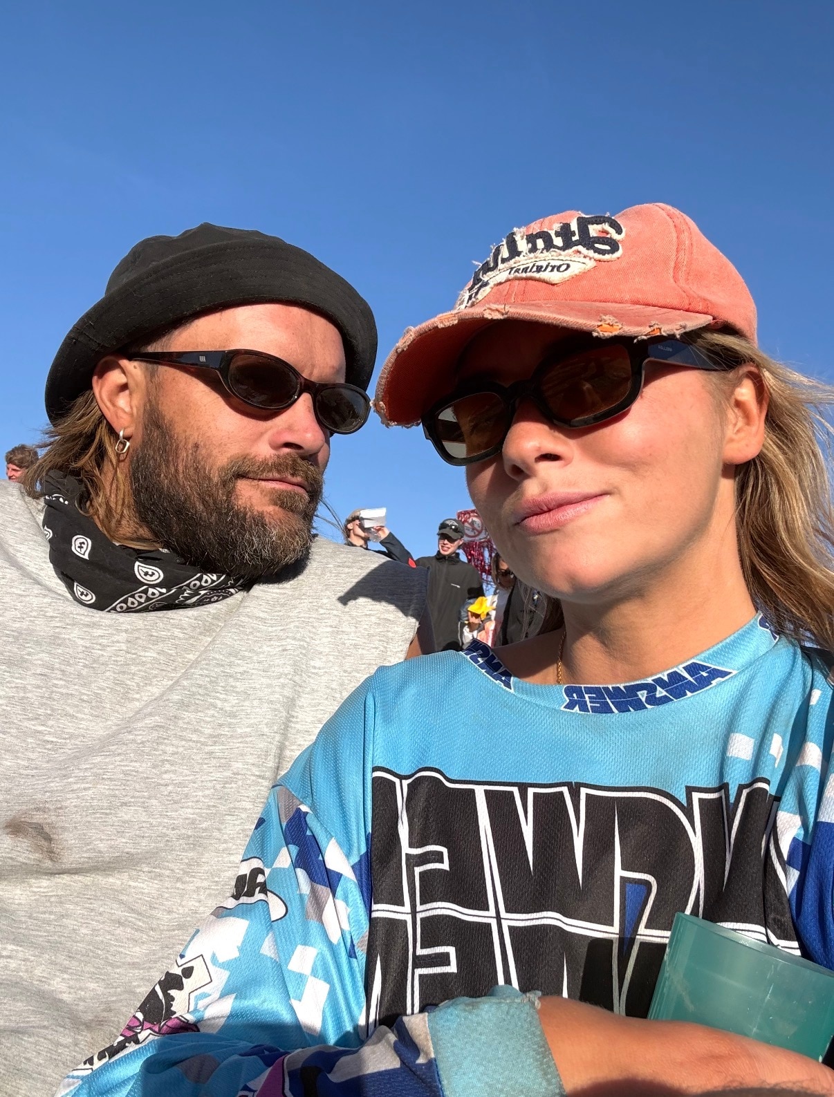 Mitch Brown de chapéu preto e óculos escuros está sentado ao lado de uma mulher loira com boné rosa e óculos escuros