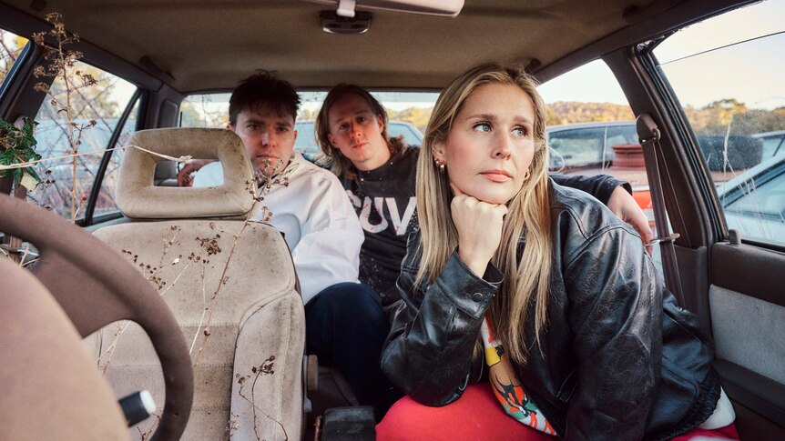 Three members of Middle Kids sit and pose in a wrecked car with foliage poking in through the windows