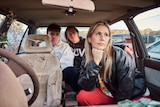 Three members of Middle Kids sit and pose in a wrecked car with foliage poking in through the windows