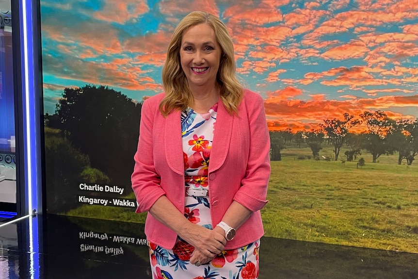 Woman standing in TV studio in front of large screen with orange clouds over green country landscape.