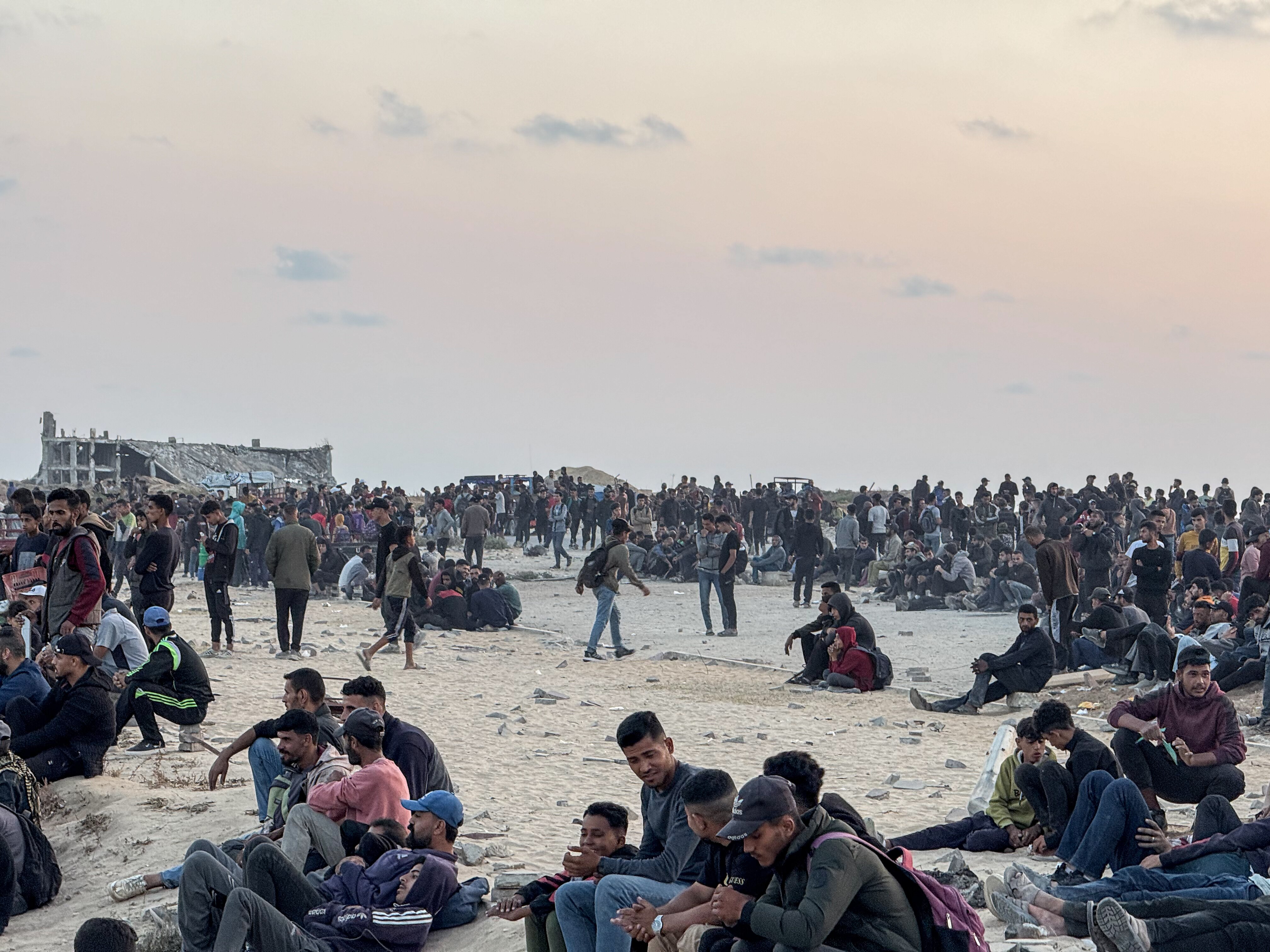 Groups of men sit, lie and stand around on barren ground with the wreckage of damaged buildings behind.