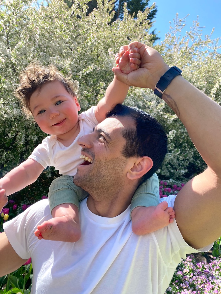 Isaac holds his smiling son on his shoulders outside on a sunny day.