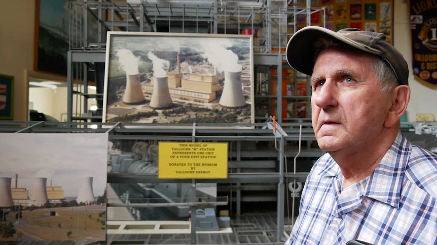 An older man wearing a cap stands in front of a picture of a power station.