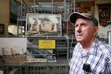 An older man wearing a cap stands in front of a picture of a power station.