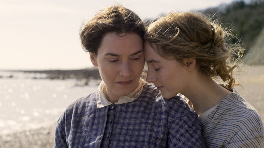 Close-up shot of Kate Winslet and Saoirse Ronan in 19th century dresses standing close together with beach in background