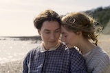 Close-up shot of Kate Winslet and Saoirse Ronan in 19th century dresses standing close together with beach in background