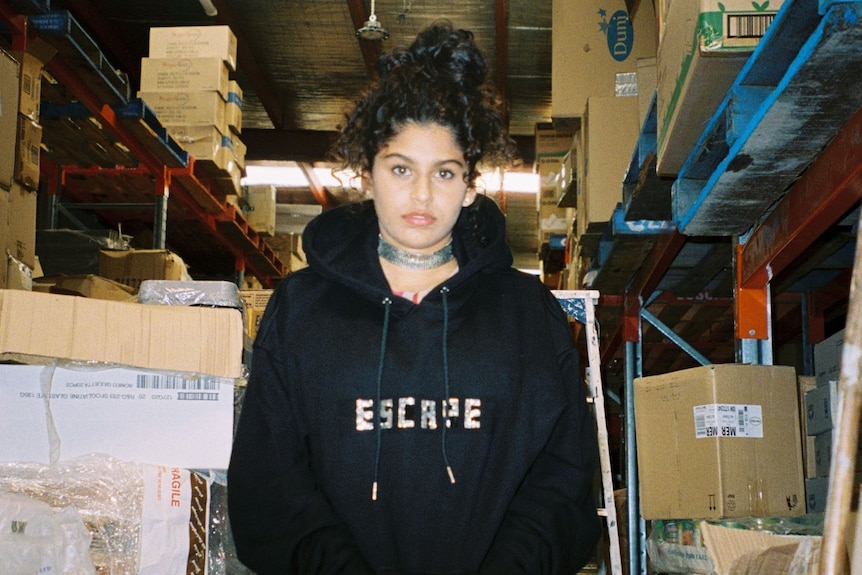 A 2022 press shot of JACOTÉNE wearing a hoodie and in a warehouse surrounded by cardboard boxes