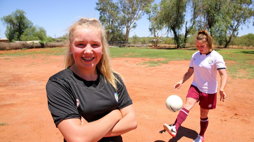 Two young girls kick around a ball on red dirt wearing their sports uniforms