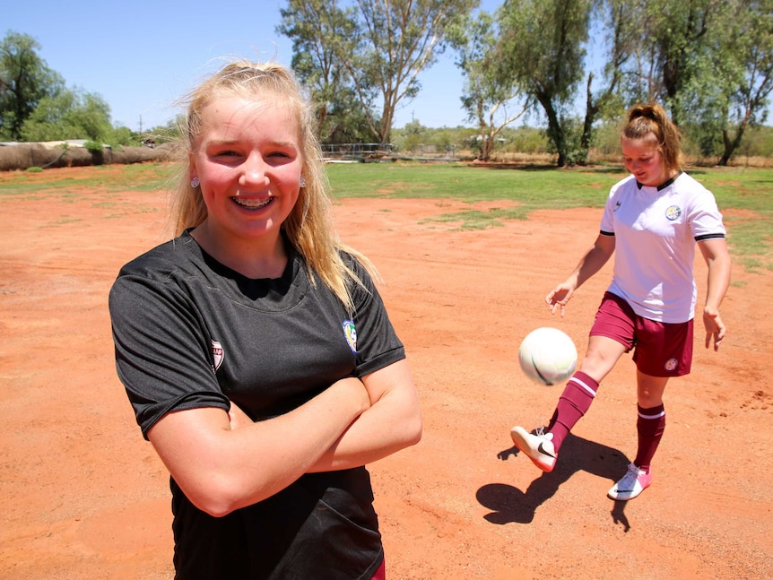 Two young girls kick around a ball on red dirt wearing their sports uniforms