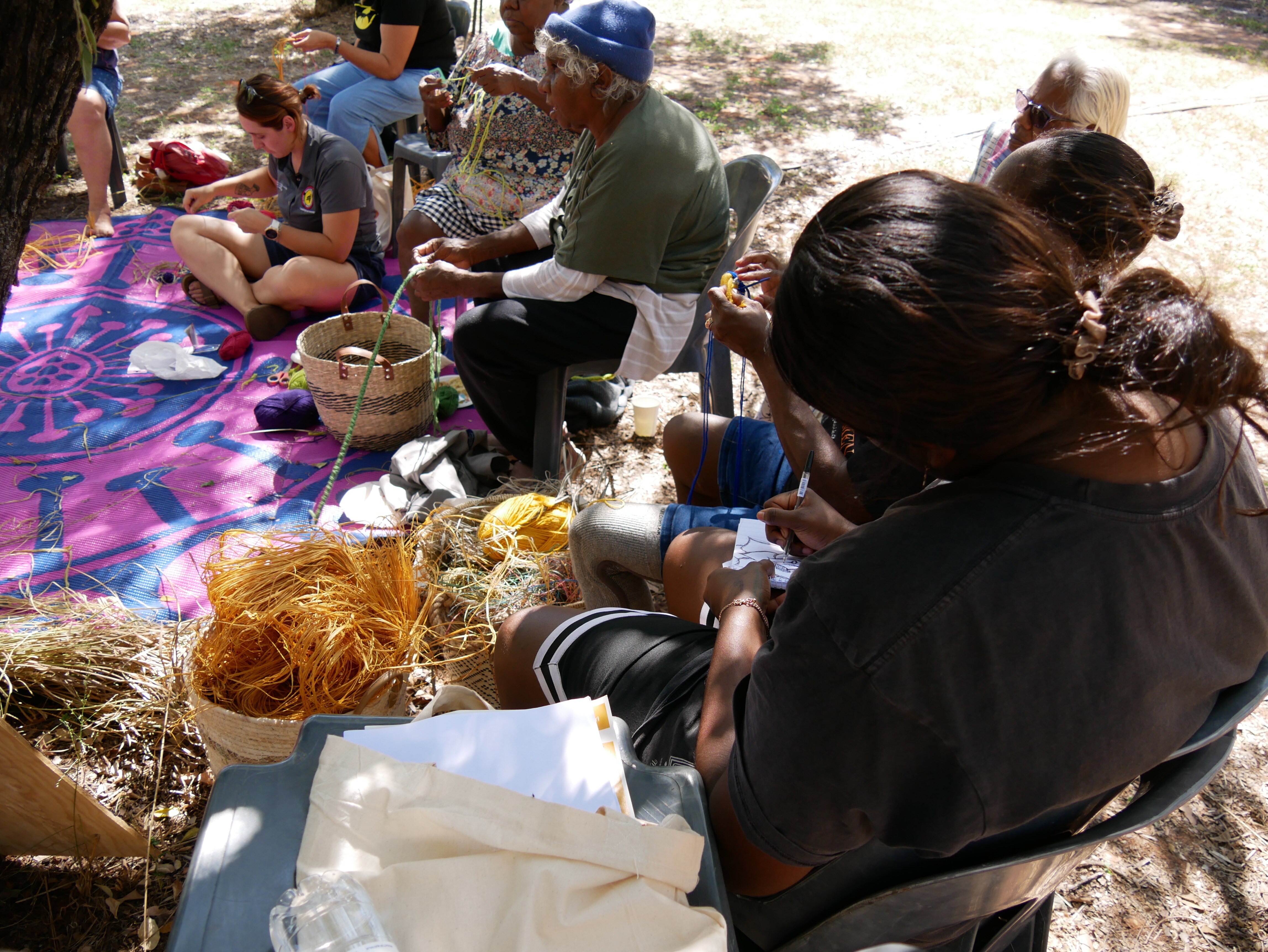Women sit around holding yarn as they weave colourful baskets