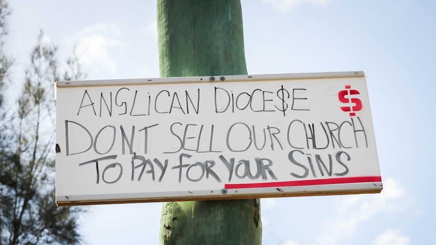 A hand written sign attached to a wooden pole that says 'Anglican Diocese Don't sell out church to pay for your sins'