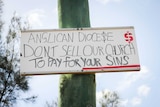A hand written sign attached to a wooden pole that says 'Anglican Diocese Don't sell out church to pay for your sins'