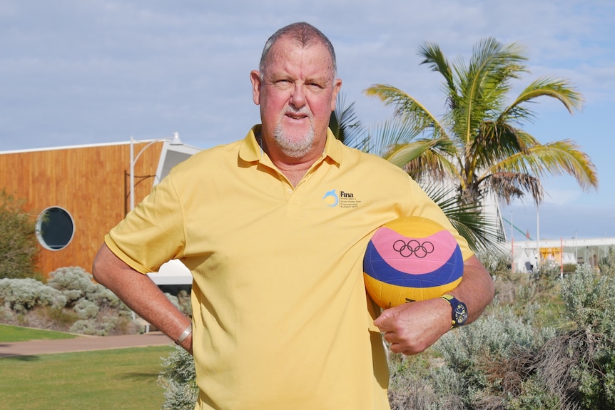 A man in a yellow polo shirt with a waterpolo ball under his arm and hand on his hip stands on grass in front of a palm tree