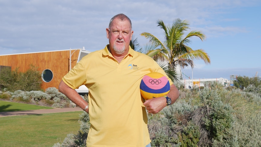 A man in a yellow polo shirt with a waterpolo ball under his arm and hand on his hip stands on grass in front of a palm tree