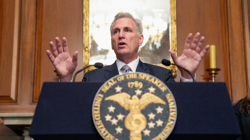 A white man in a suit stands and gestures at a us government podium 