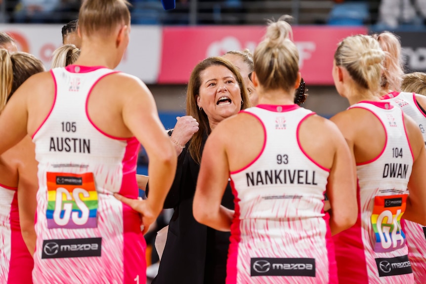 A Super Netball coach gestures as she speaks to a circle of her players during a match.