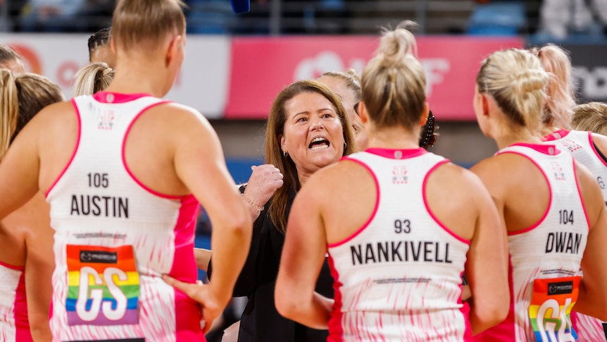 A Super Netball coach gestures as she speaks to a circle of her players during a match.