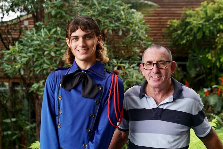 Tall trans caucasian woman wearing a blue military-style dress stands next to her short caucasian father.