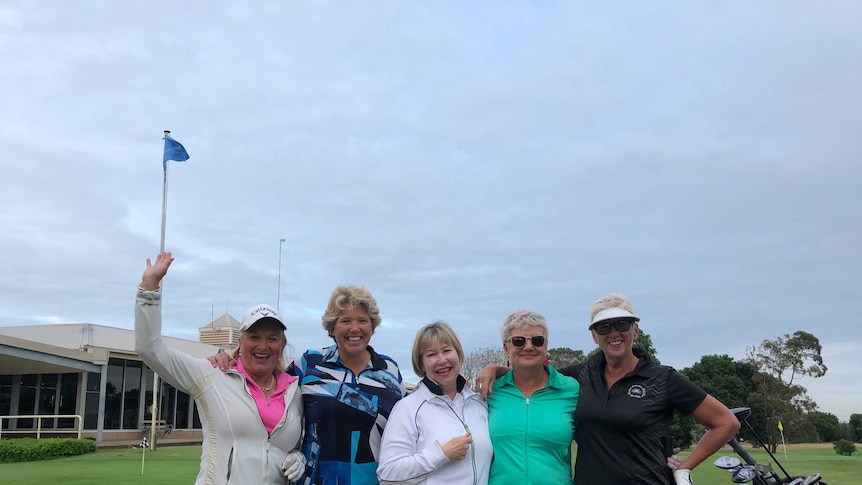 A group of five women in golf outfits standing in a group at a golf course and cheering.